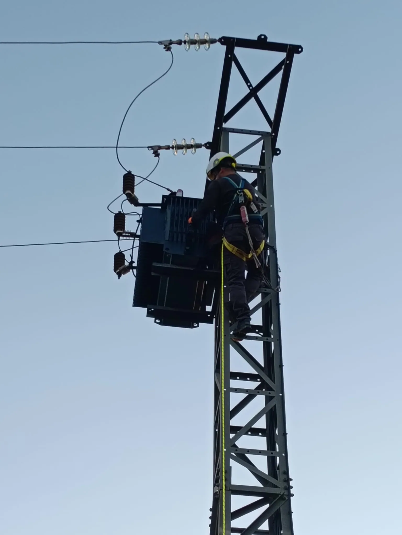 Electricista con arnés de seguridad realizando montaje de transformador en apoyo eléctrico en altura
